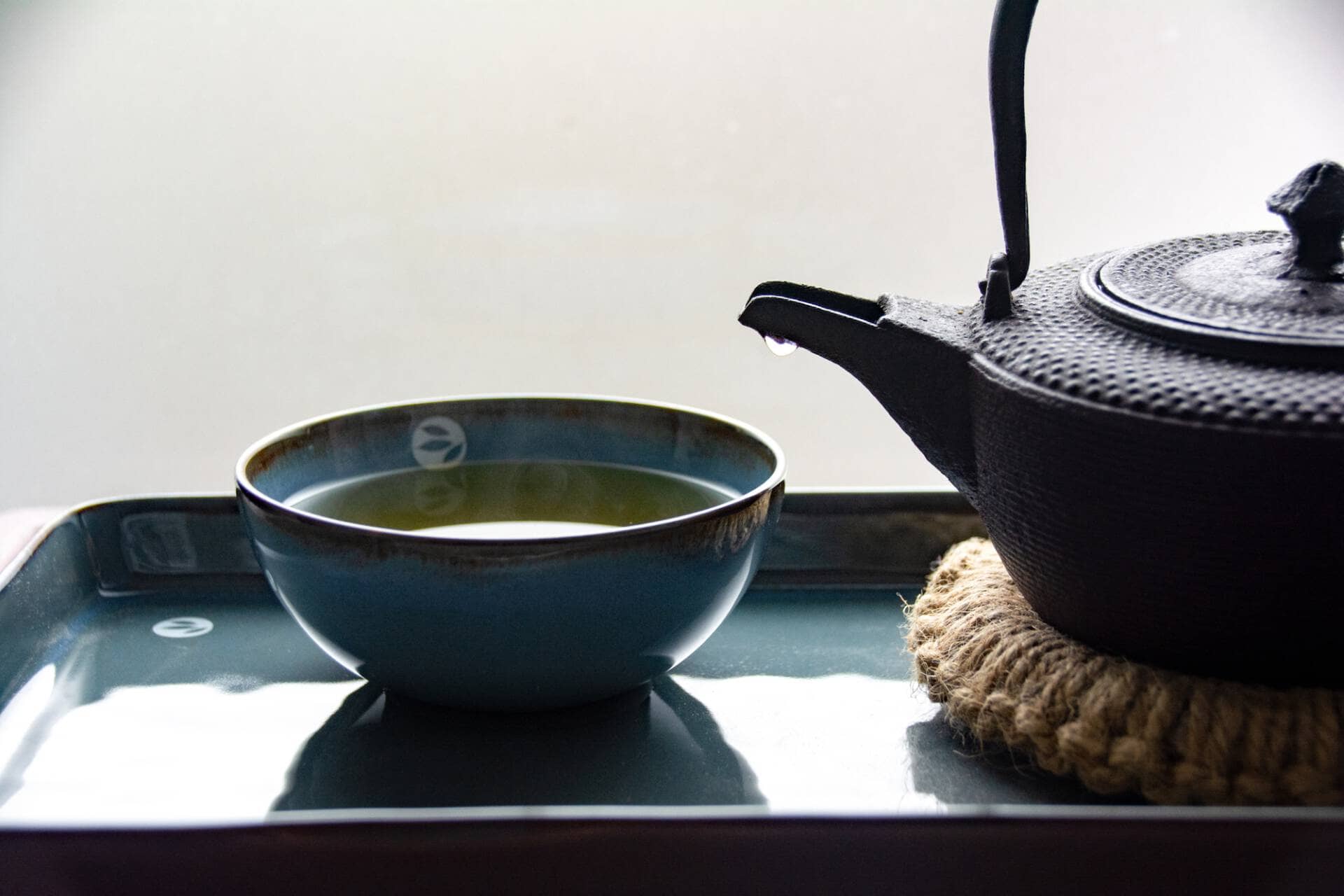 Tea_Passion_Sven-Christian Lange_Branding Photography_Ancient Black Cast Iron Kettle On Coaster Made Of Bast With Drop On Spout And Steaming Turquoise Tea Bowl On Platter