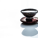 Tea Passion_Sven-Christian Lange_Branding Photography_Beautiful Antique Black Korean Pottery Art Bowl On Wooden Coaster With Little Tempelflowers_On White Reflecting Surface_Green Tea__Tea Ceremony