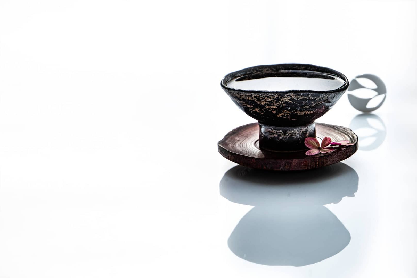 Tea Passion_Sven-Christian Lange_Branding Photography_Beautiful Antique Black Korean Pottery Art Bowl On Wooden Coaster With Little Tempelflowers_On White Reflecting Surface_Green Tea__Tea Ceremony